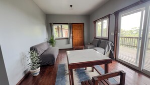 Une pièce avec un plancher en bois, une table blanche, un canapé gris, un banc en bois, une plante en pot et une vue sur un balcon à travers une grande fenêtre.
