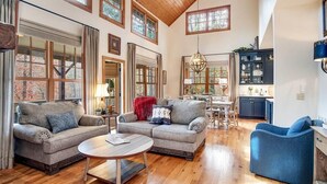 A spacious living room with a high ceiling, wooden floors, and large windows. There are two sofas, a coffee table, and a dining area with a chandelier. - Fern Cliff - A cozy retreat in Cashiers (Cashiers)