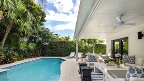 A poolside area with a curved pool, lounge chairs, a sofa, and a ceiling fan.