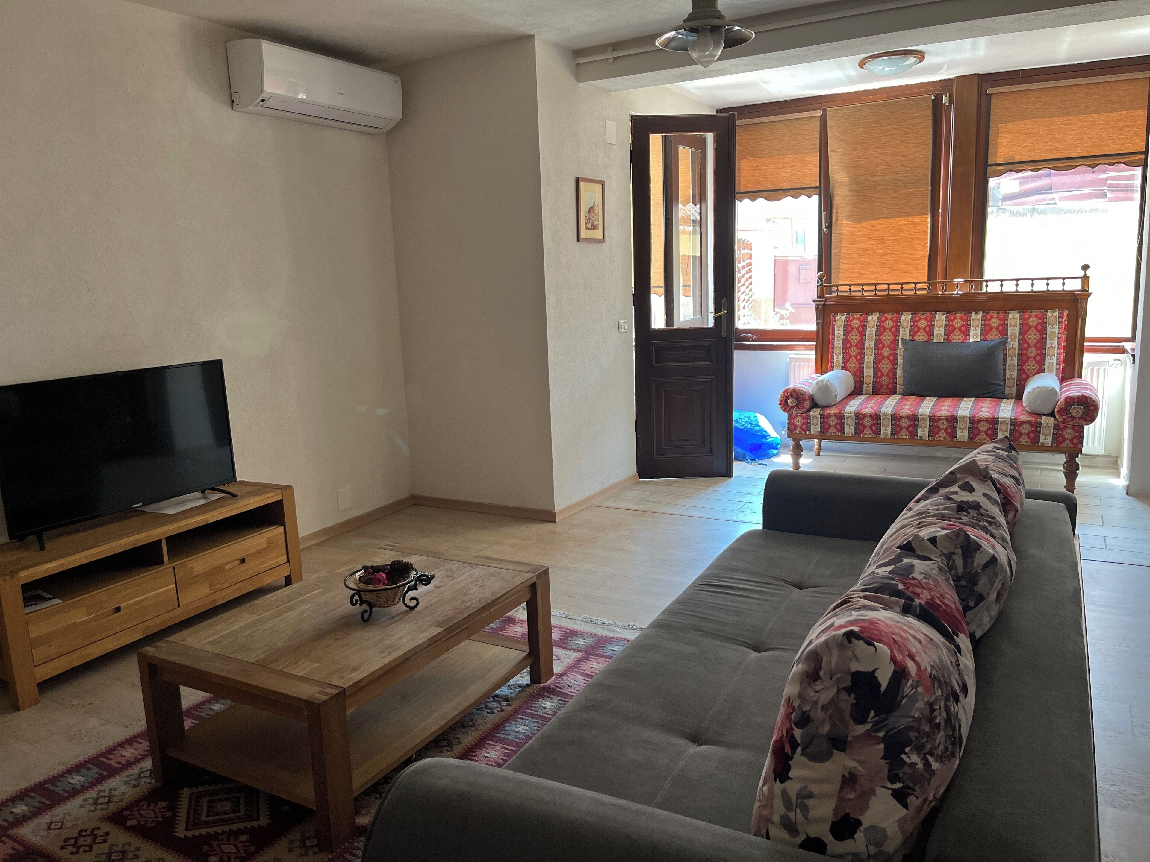Ein Wohnzimmer mit Fernseher auf Holzfuß, einer Couch mit floralen Kissen, einem Holztisch und einem gemusterten Teppich.