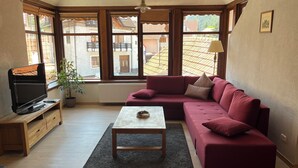 A cozy living room with a sectional sofa, a coffee table, a TV on a wooden stand, and large windows offering a view of buildings and trees. - Cozy Apartment (Brasov)