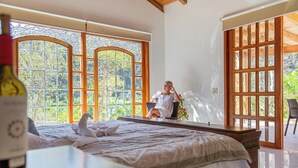 A bedroom with a large bed, a window with a view of greenery, and a bottle of wine on the bedside table. -  La Dulce Vista Hotel & Villas - Sierra Madre Puerto Vallarta (Puerto Vallarta)