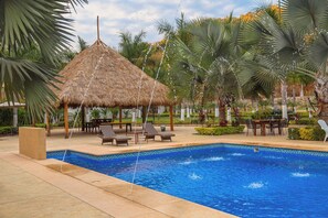 A pool area with a thatched-roof structure, lounge chairs, and palm trees.