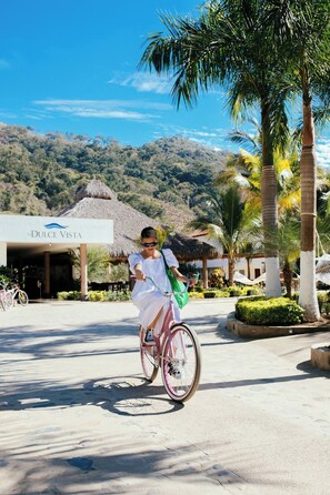 A person riding a pink bicycle in front of a resort named "La Dulce Vista" with a thatched-roof building and palm trees.