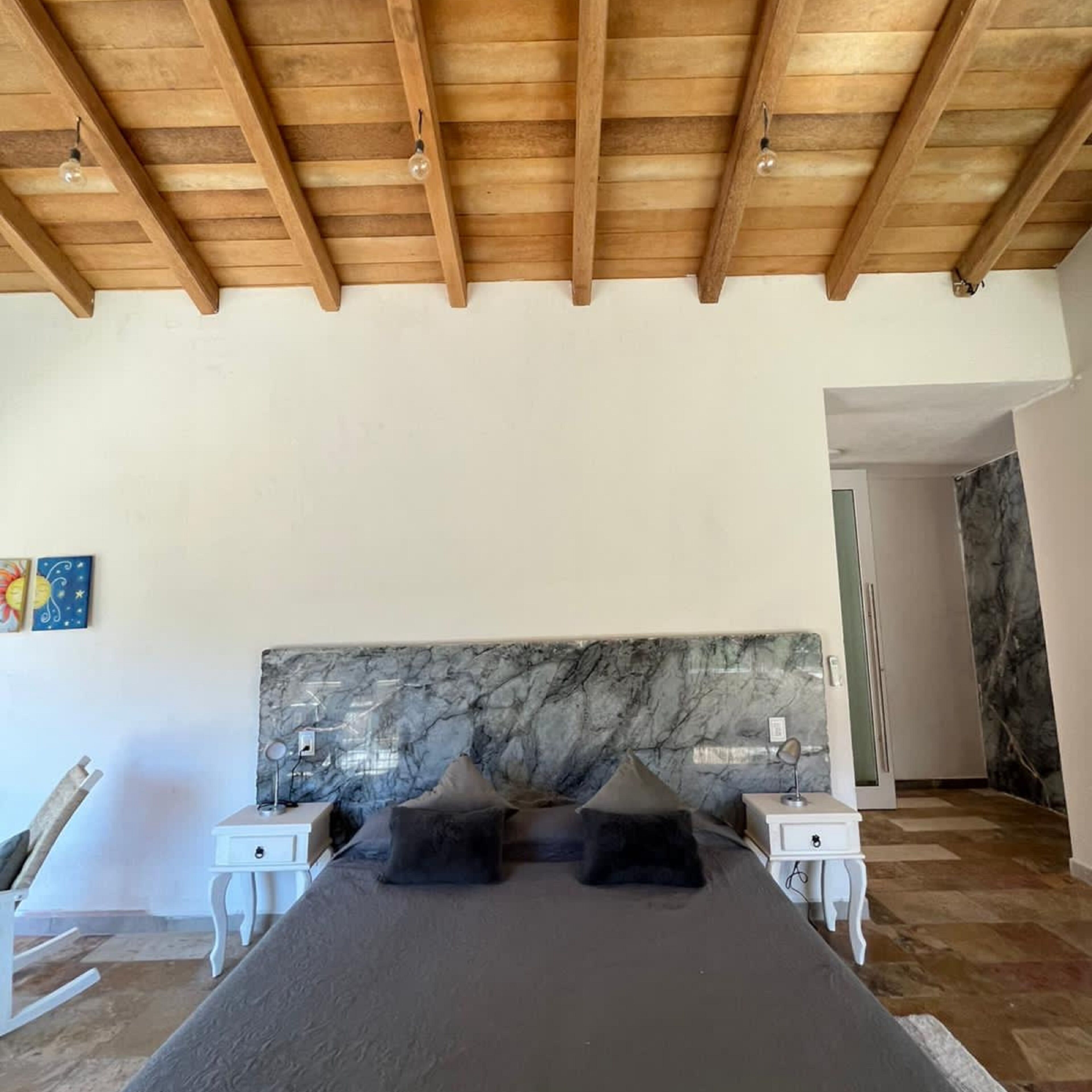 A bedroom with a large bed, marble headboard, wooden ceiling beams, and white nightstands.