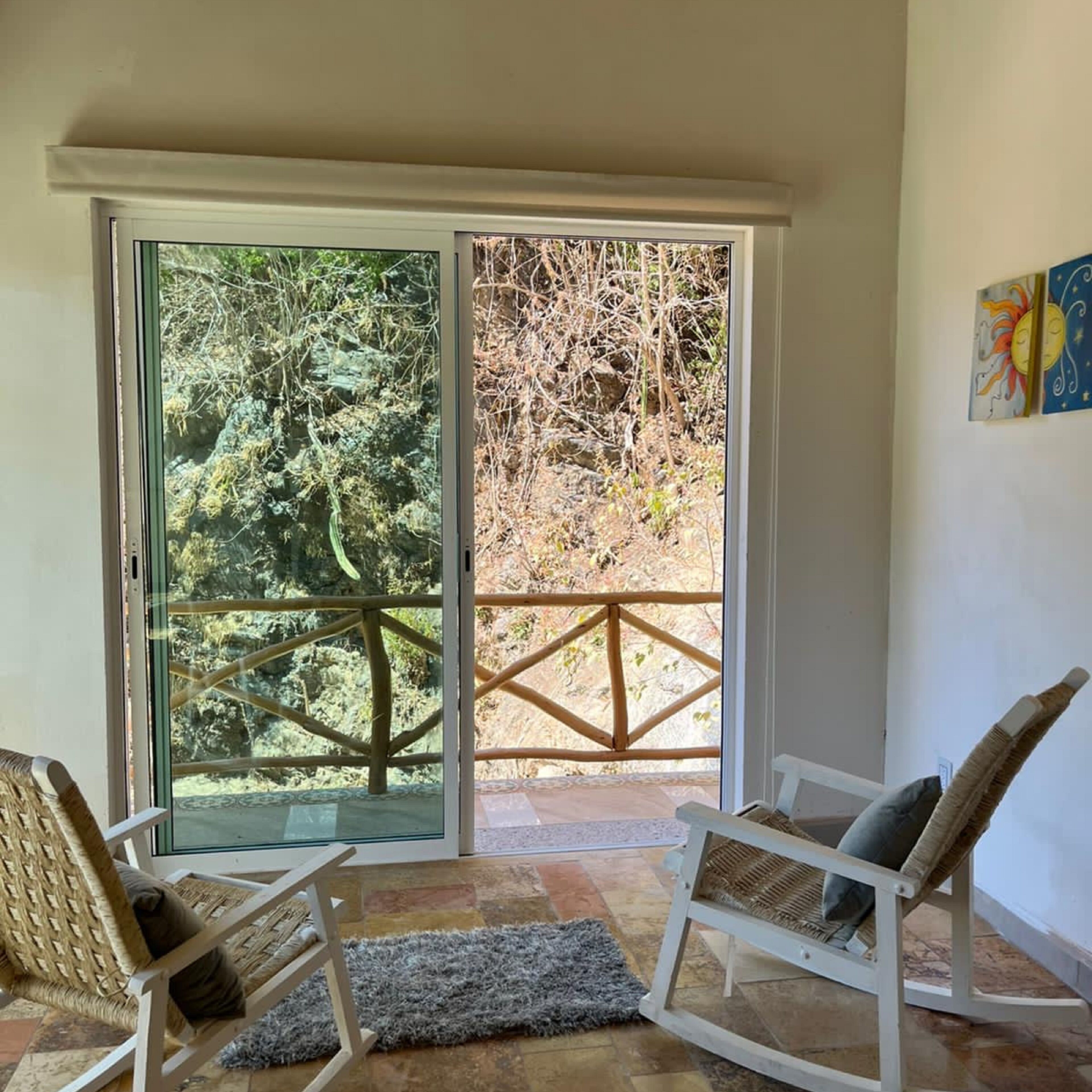 Una habitación con dos sillas de mimbre, un tapete gris y un gran ventanal corredizo que da a un balcón con barandilla de madera y vistas a árboles.