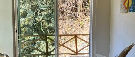 Una habitación con dos sillas de mimbre, un tapete gris y un gran ventanal corredizo que da a un balcón con barandilla de madera y vistas a árboles.
