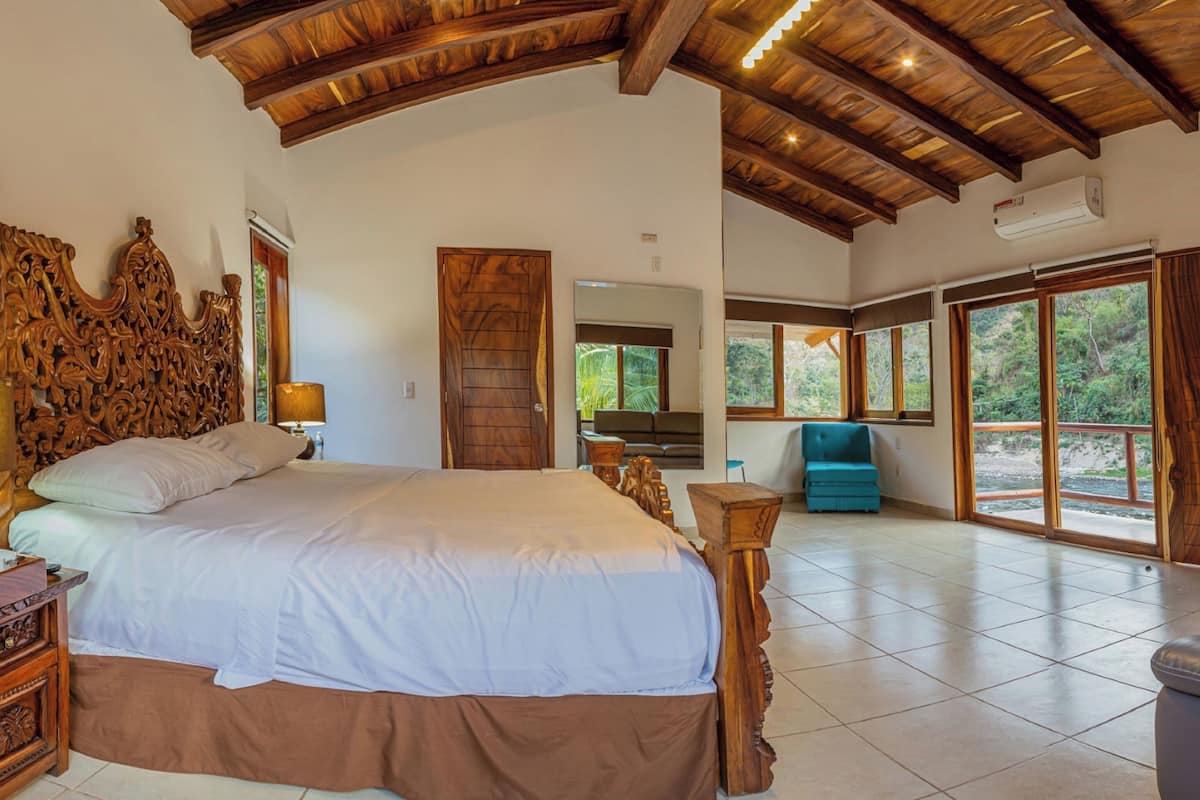 A spacious bedroom with a large bed, wooden headboard, and a view of the outdoors through large windows.