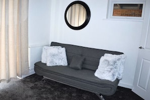 A grey sofa with two white fluffy cushions, a round mirror on the wall, and a door with a window above it.
