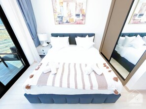 A modern bedroom with a large bed, a bedside table with a lamp, and a view of the outdoors. - Wonderful 2B With Breathtaking View in JVC (Dubai)