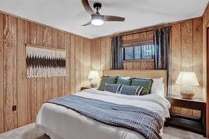 A bedroom with a wooden paneled wall, a bed with white bedding and blue accents, two bedside lamps, a ceiling fan, and a window with curtains. - Winter Getaway | Hot Tub & Sauna | Game Room (Clifton)