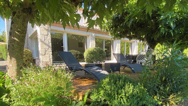 Un área de estar al aire libre con techado, sillones reclinables y una pequeña mesa, rodeada de exuberante follaje y un edificio de piedra al fondo.