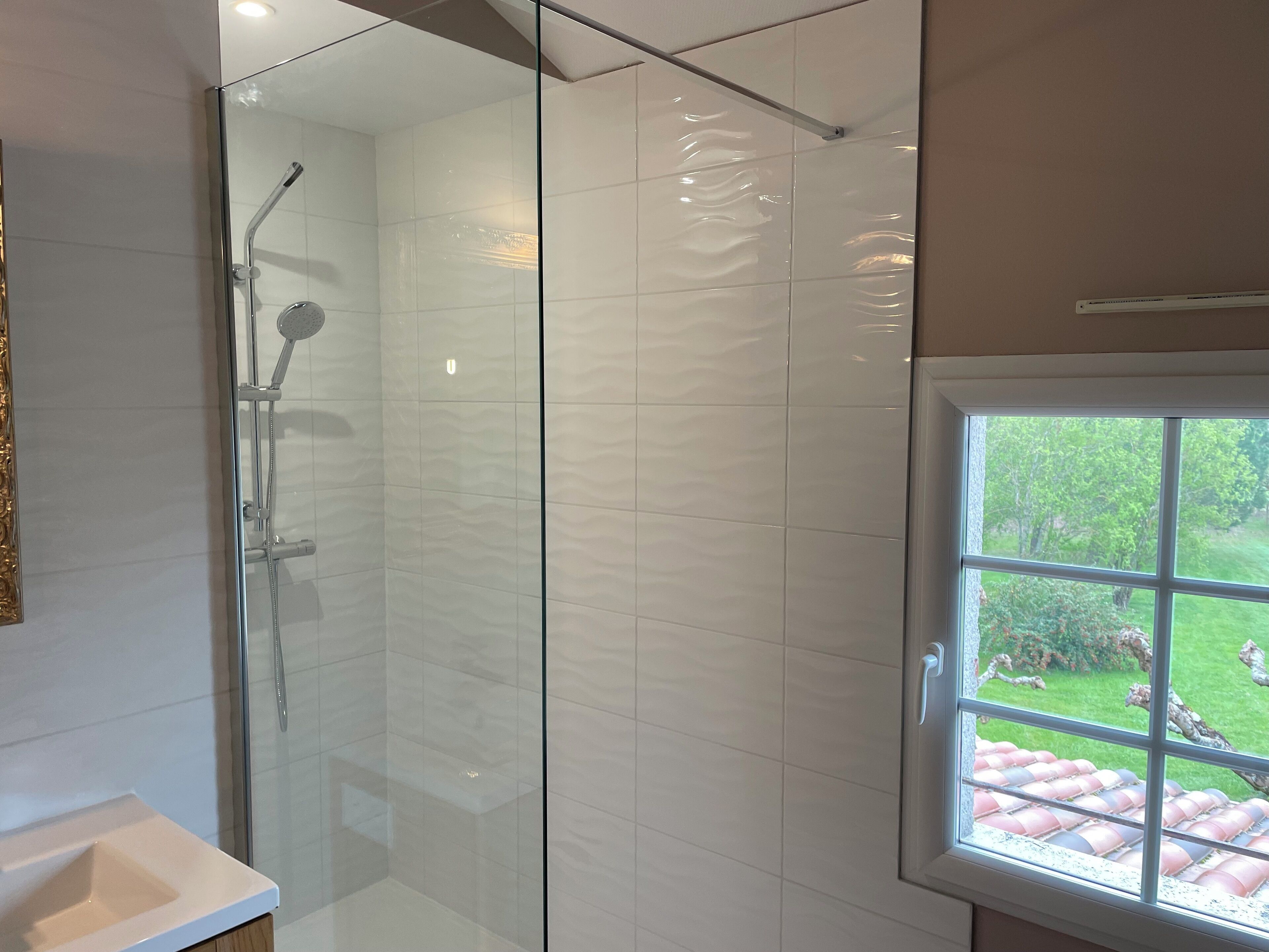 A modern bathroom with a glass-enclosed shower, a white sink, and a window offering a view of a garden.