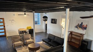 Ett modernt vardagsrum med en svart lädersoffa, ett runt soffbord och ett sidobord i trä. I taket hänger en takfläkt, på väggen finns en darttavla och en målning som föreställer en stadsbild.