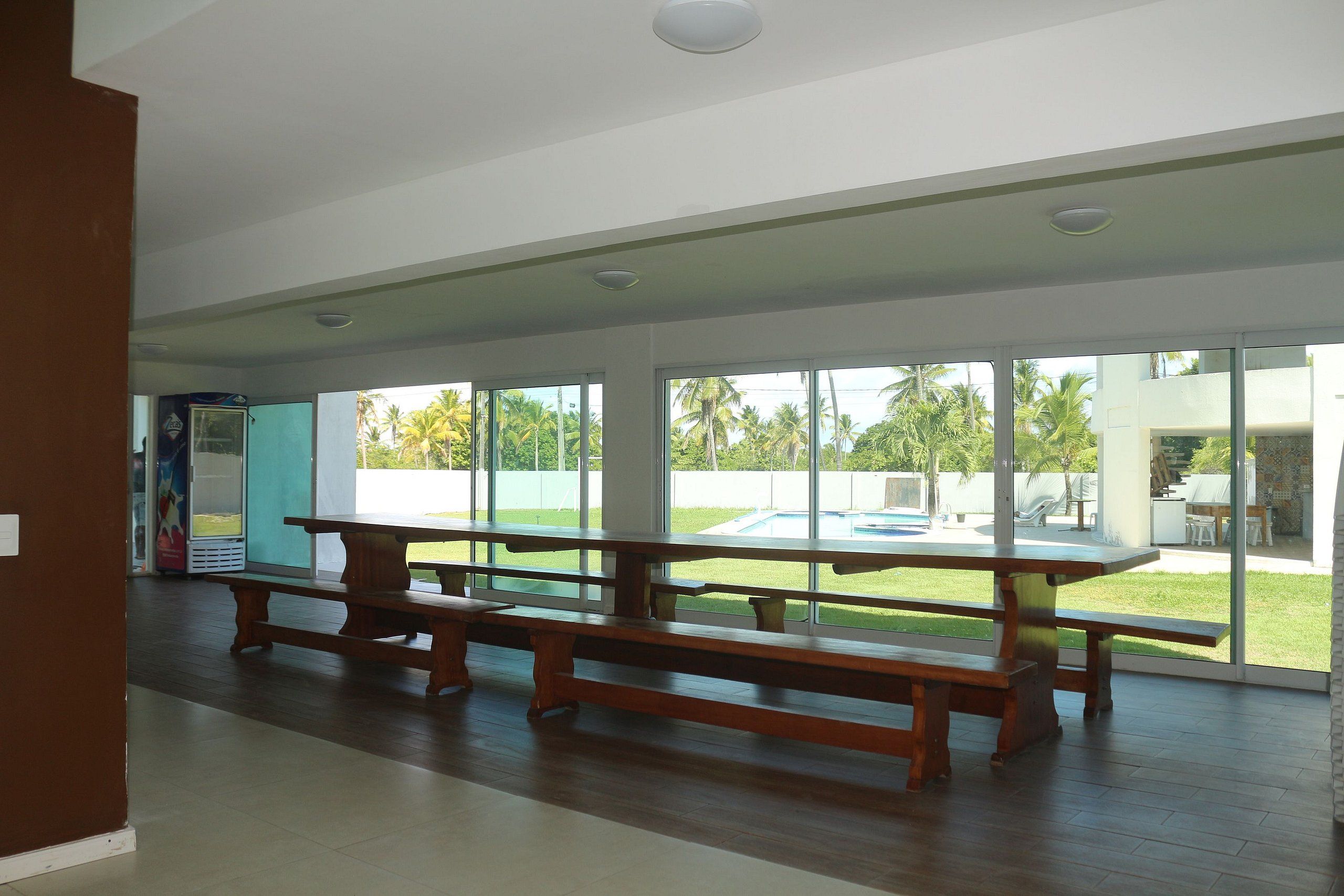 Amplo espaço interno com uma grande mesa de madeira e bancos, janelas grandes com vista para uma piscina e palmeiras.