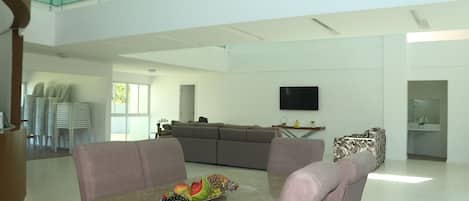 A spacious living area with a dining table set with fruit, a grey sofa, and a television mounted on the wall.