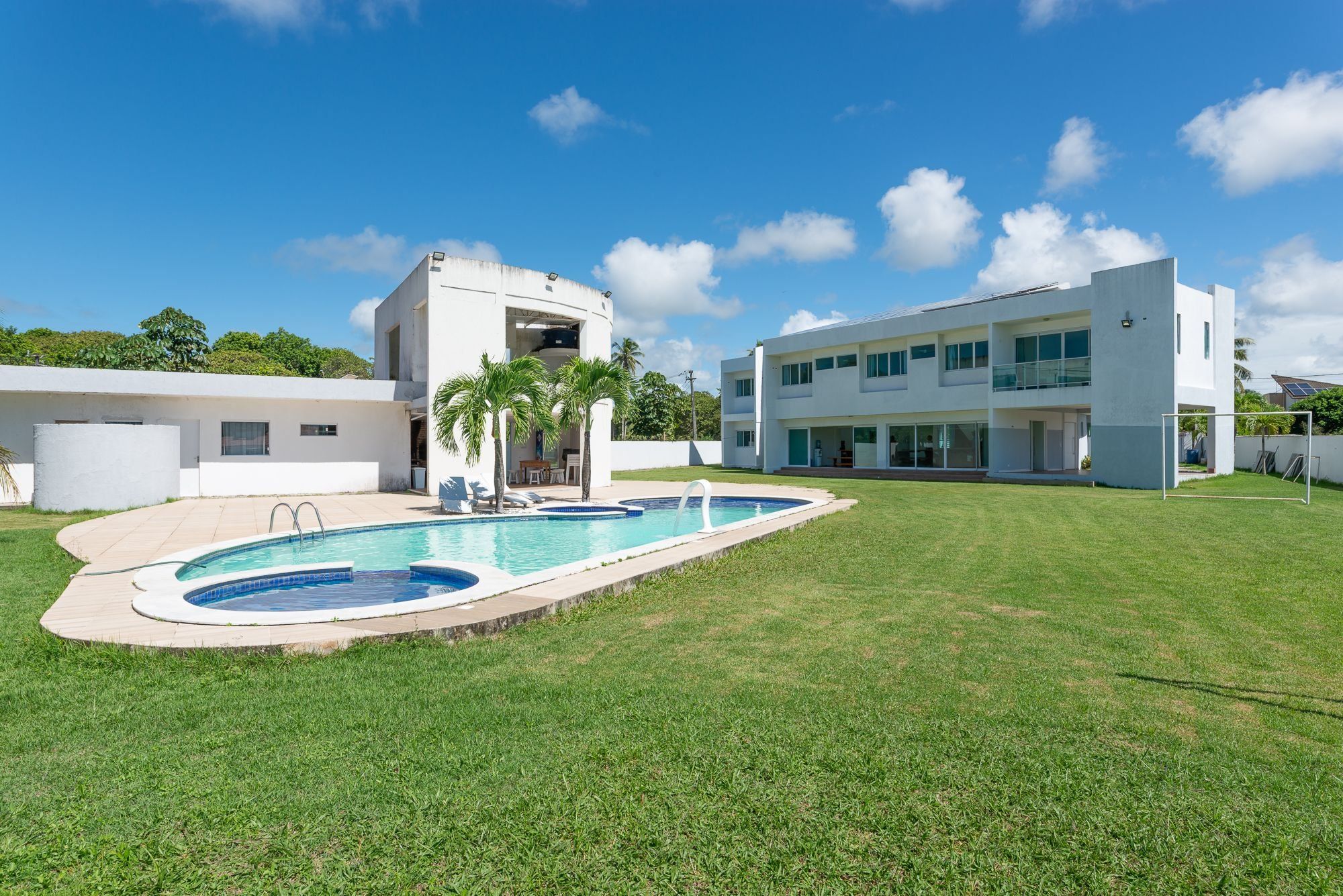 Un edificio moderno de dos pisos con piscina, palmeras y un cielo azul despejado.