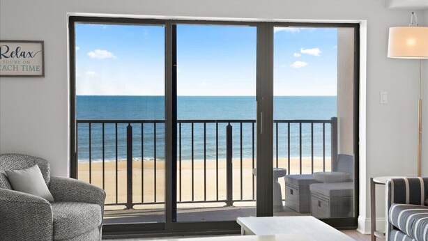 A room with a large sliding glass door leading to a balcony with a view of the beach. There is a sofa and a table in the room. - Luxe Oceanfront 1BR | Pool, Hot Tub, Sun Deck (Ocean City)