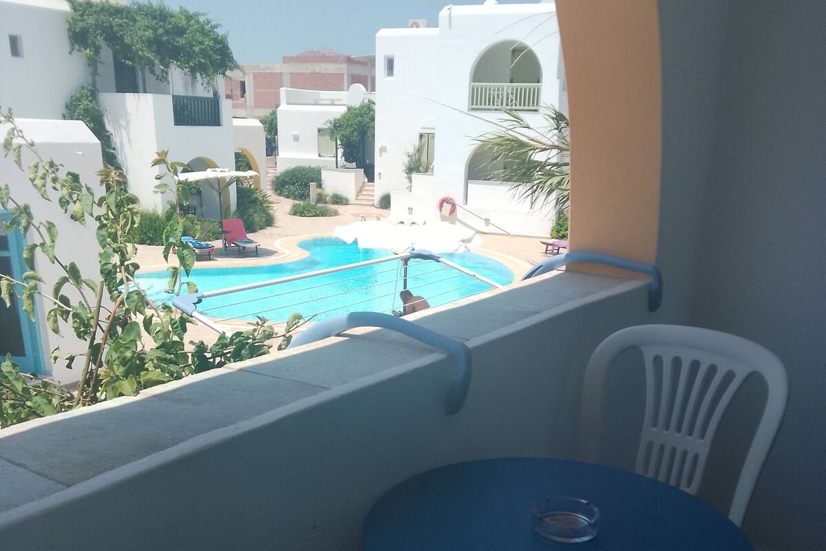 A balcony with a pool view, white chairs, and a small table.