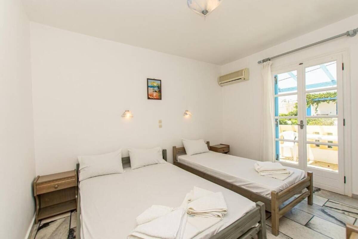 A room with two single beds, a small table, a wall-mounted air conditioner, and a window with a view of the outside.