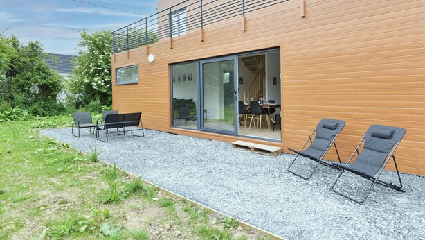 Ein modernes Gebäude mit Holzfassade, einer überdachten Sitzgelegenheit im Freien mit zwei Liegestühlen und einer Kiesfläche.