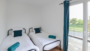 Ein kleiner, sauberer Raum mit zwei Einzelbetten, jeweils mit weißer Matratze und türkisfarbenen Kissen. Ein Fenster mit türkisblauem Vorhang, und durch das Fenster ist ein Balkon mit Metallgeländer zu sehen.