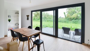 Un comedor con una mesa y sillas de madera, una estantería blanca con cuadros enmarcados, y un amplio ventanal corredera que da a un jardín.
