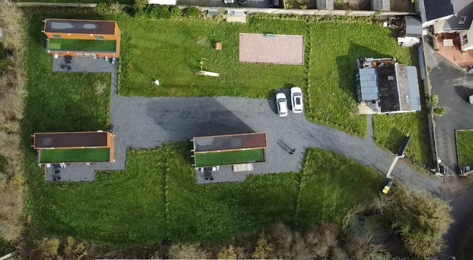 An aerial view of a property with three green-roofed buildings, a paved driveway, and two parked cars.
