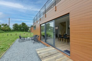 Une maison moderne avec une façade en bois, un balcon doté de rampes en métal et une terrasse aménagée avec du mobilier d’extérieur.