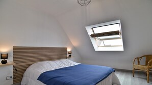 Ein Schlafzimmer mit schräg verlaufender Decke, einem Bett mit weiß-blauer Bettdecke, einem Holzkopfende, einem kleinen Nachttisch mit Lampe, einem Korbstuhl und einem Fenster mit Klimaanlage.