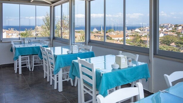 A dining area with tables and chairs, large windows with a view of a coastal town and a bridge. - Begonvil Hotel Bozcaada (Bozcaada)