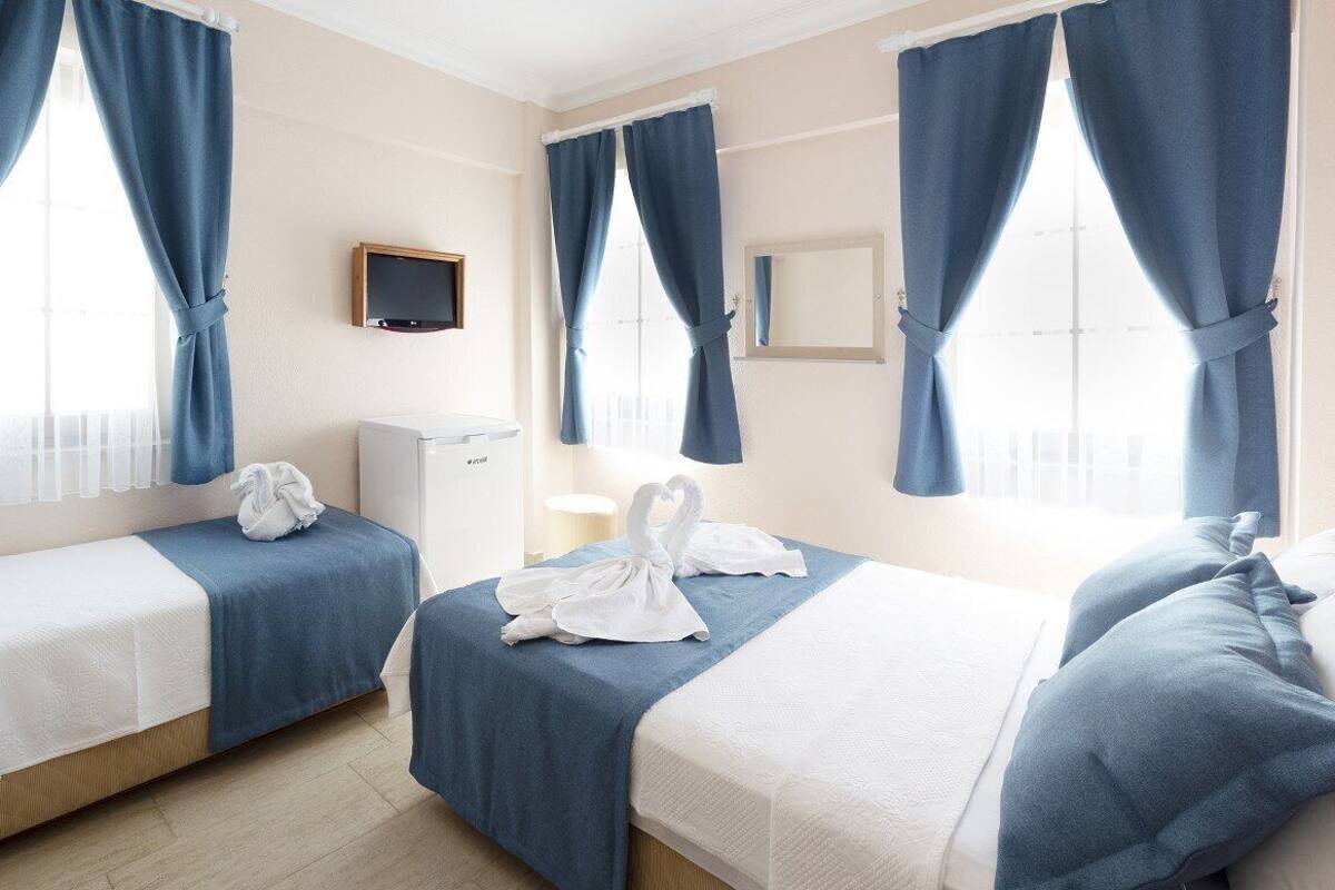 A hotel room with two beds, each with a blue bedspread and white pillows. There is a flat-screen TV mounted on the wall, a small table, and a window with blue curtains.