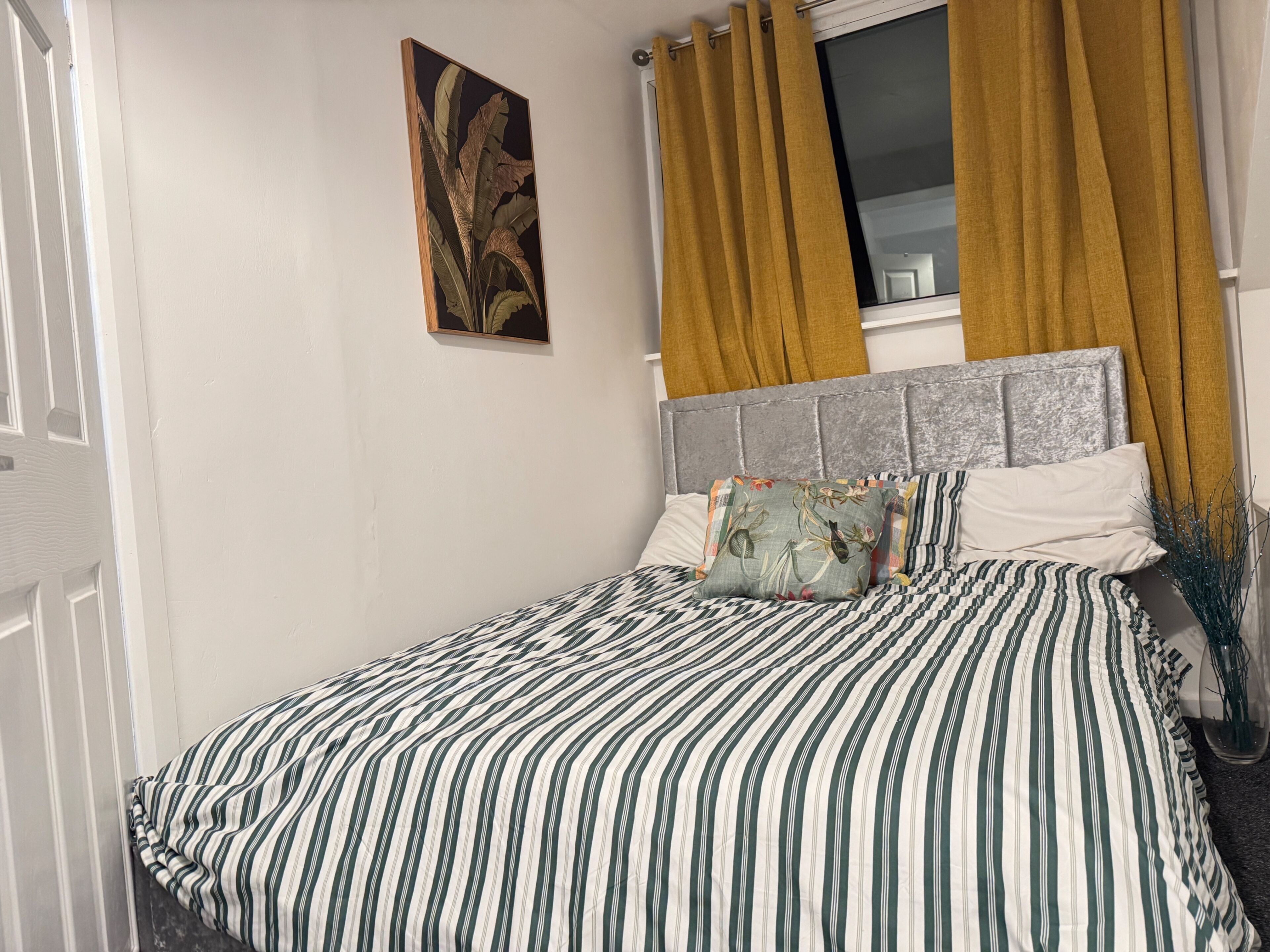 A bedroom with a striped bedspread, a grey headboard, and a window with yellow curtains.