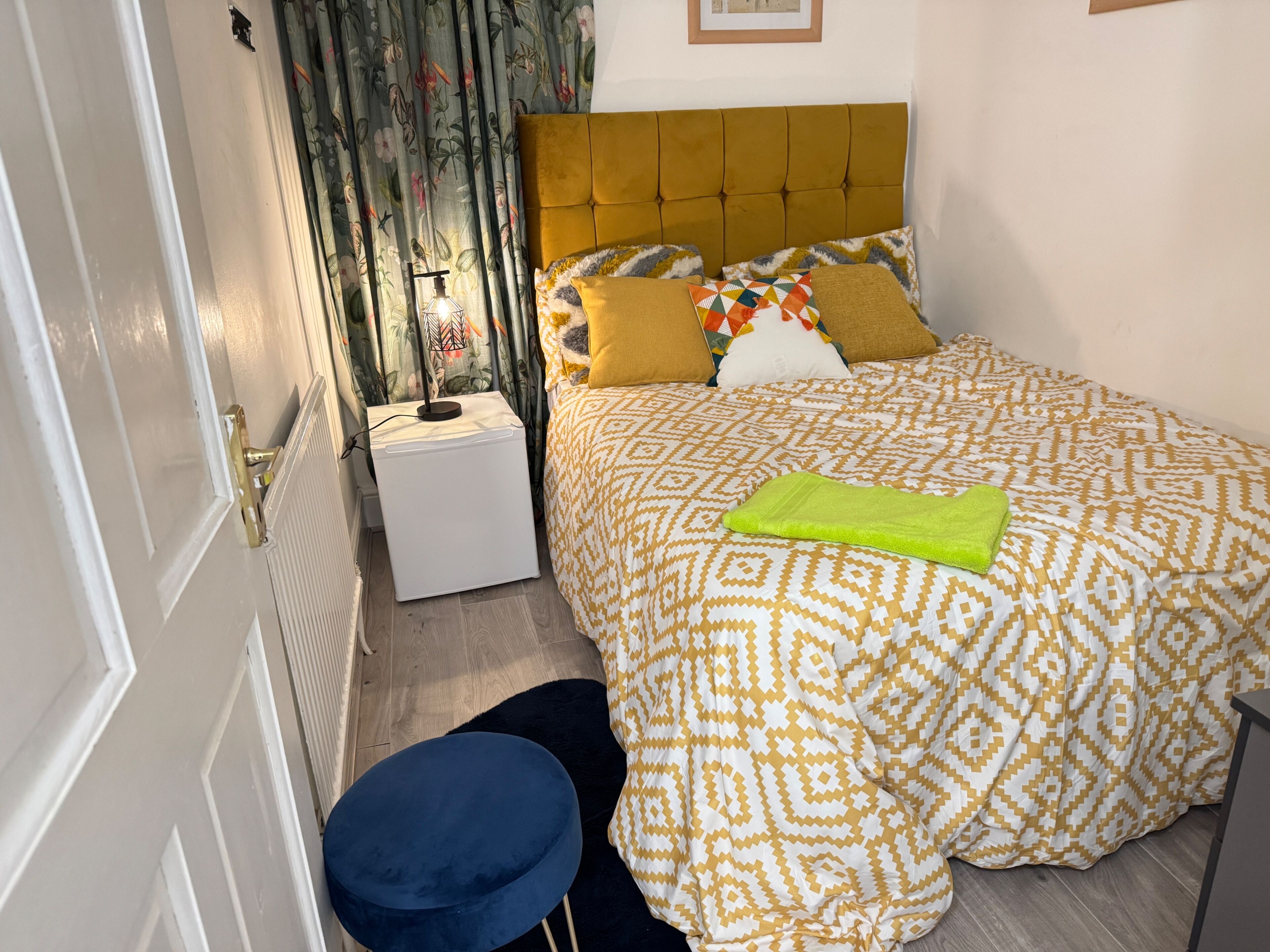 A neatly arranged bedroom with a patterned bedspread, a bedside lamp, and a small round stool.