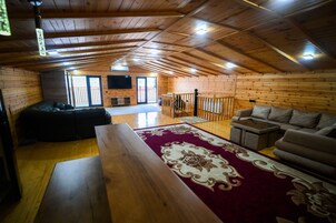 Interior acolhedor de uma cabana com teto de madeira, uma janela grande, sofá, mesa de centro e televisão.