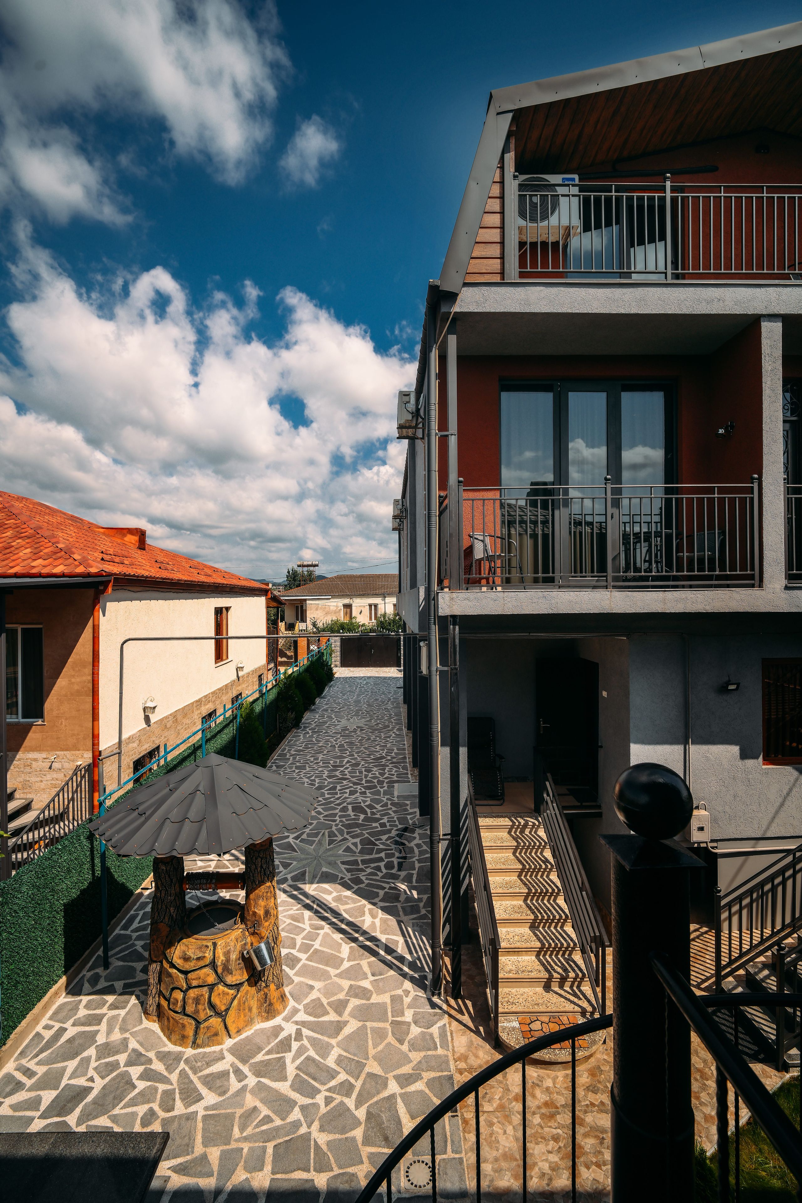 A courtyard with a stone floor, a tree with a metal bucket, and a building with a balcony.