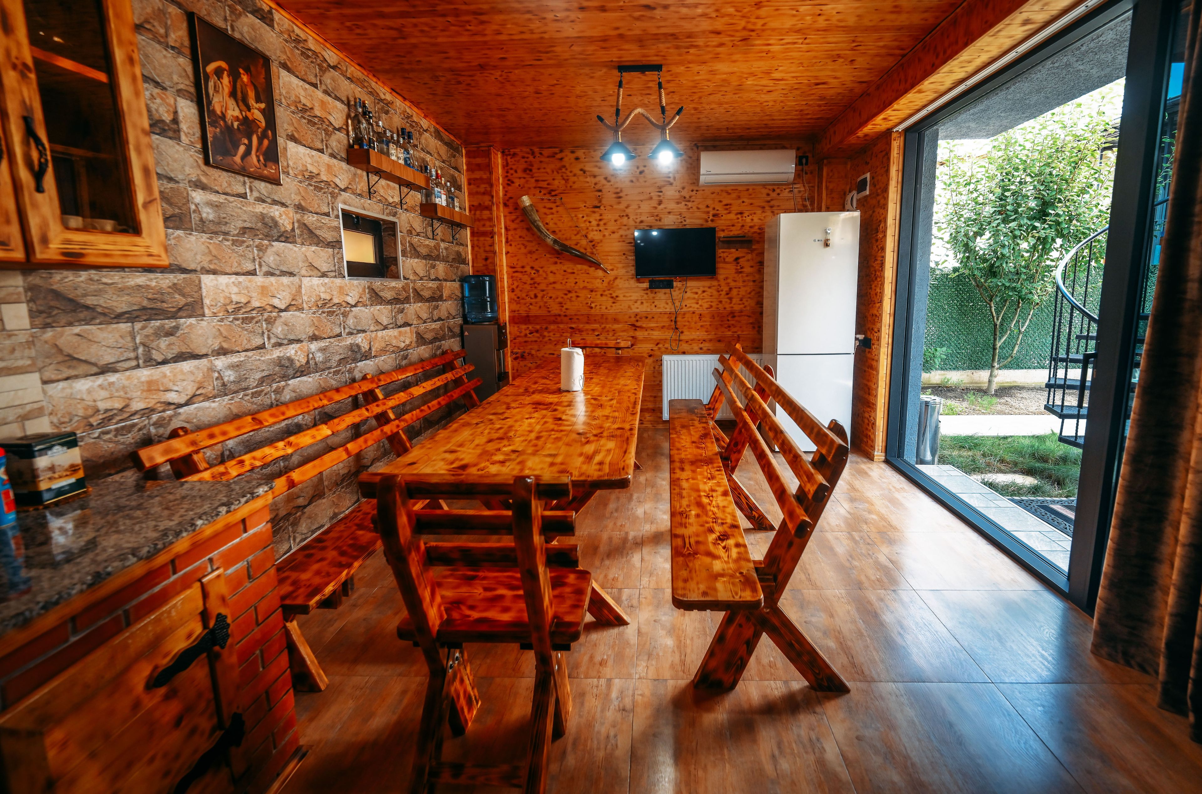 A wooden interior with a long dining table, benches, stone walls, and a view through a glass door.