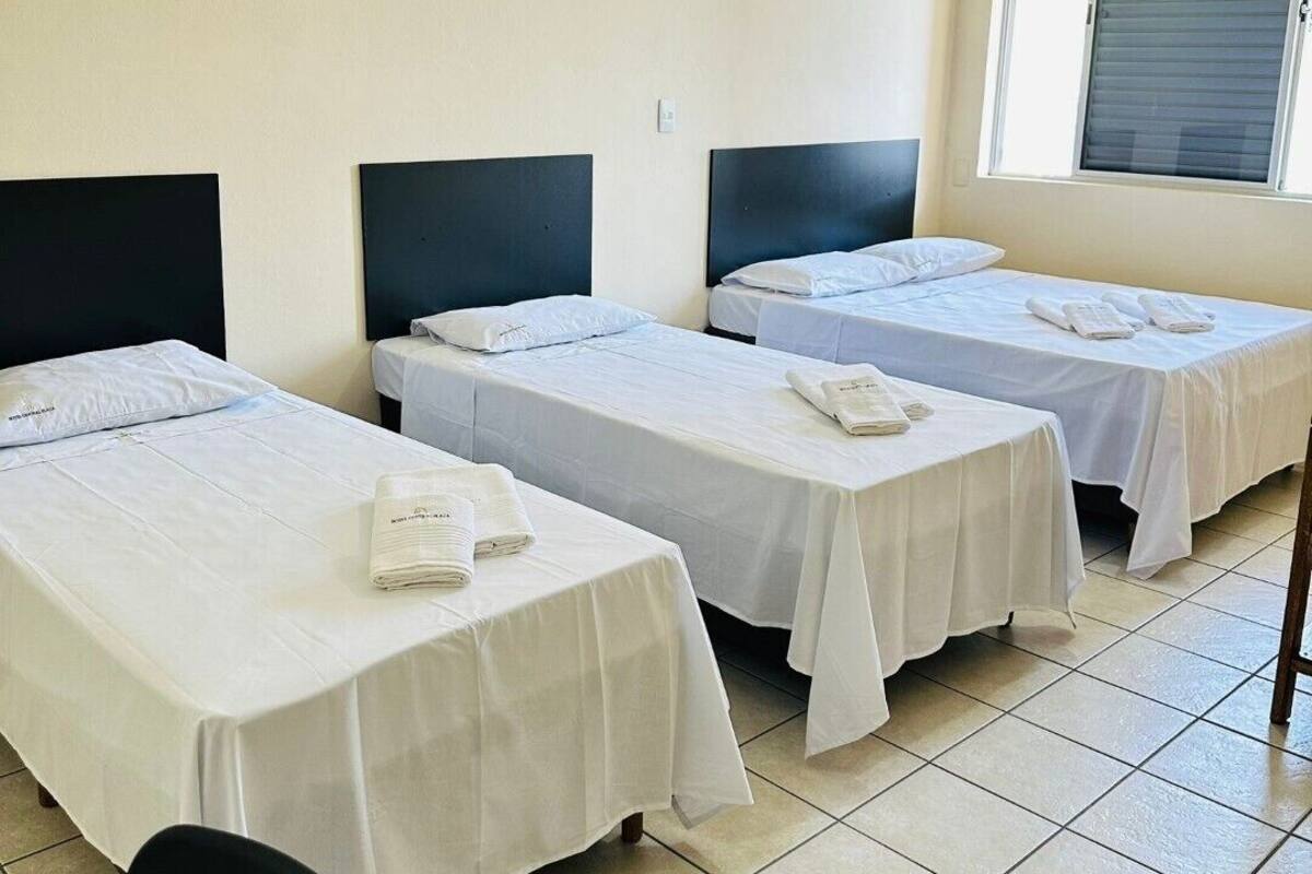 A hotel room with three single beds, each with a black headboard and white bedding. There is a window with blinds, a chair, and a table with a folded towel on it.