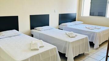 Camera d'albergo con tre letti singoli, ognuno dotato di una testiera nera e biancheria da letto bianca. Si nota una finestra con persiane, una sedia e un tavolo con un asciugamano piegato.