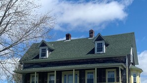 A two-story house with a green roof, a porch with white railings, and a small front yard with grass and dandelions.