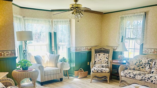 A cozy living room with a wooden floor, a ceiling fan, and two windows with curtains. There are two sofas, a coffee table, and a side table with a lamp.