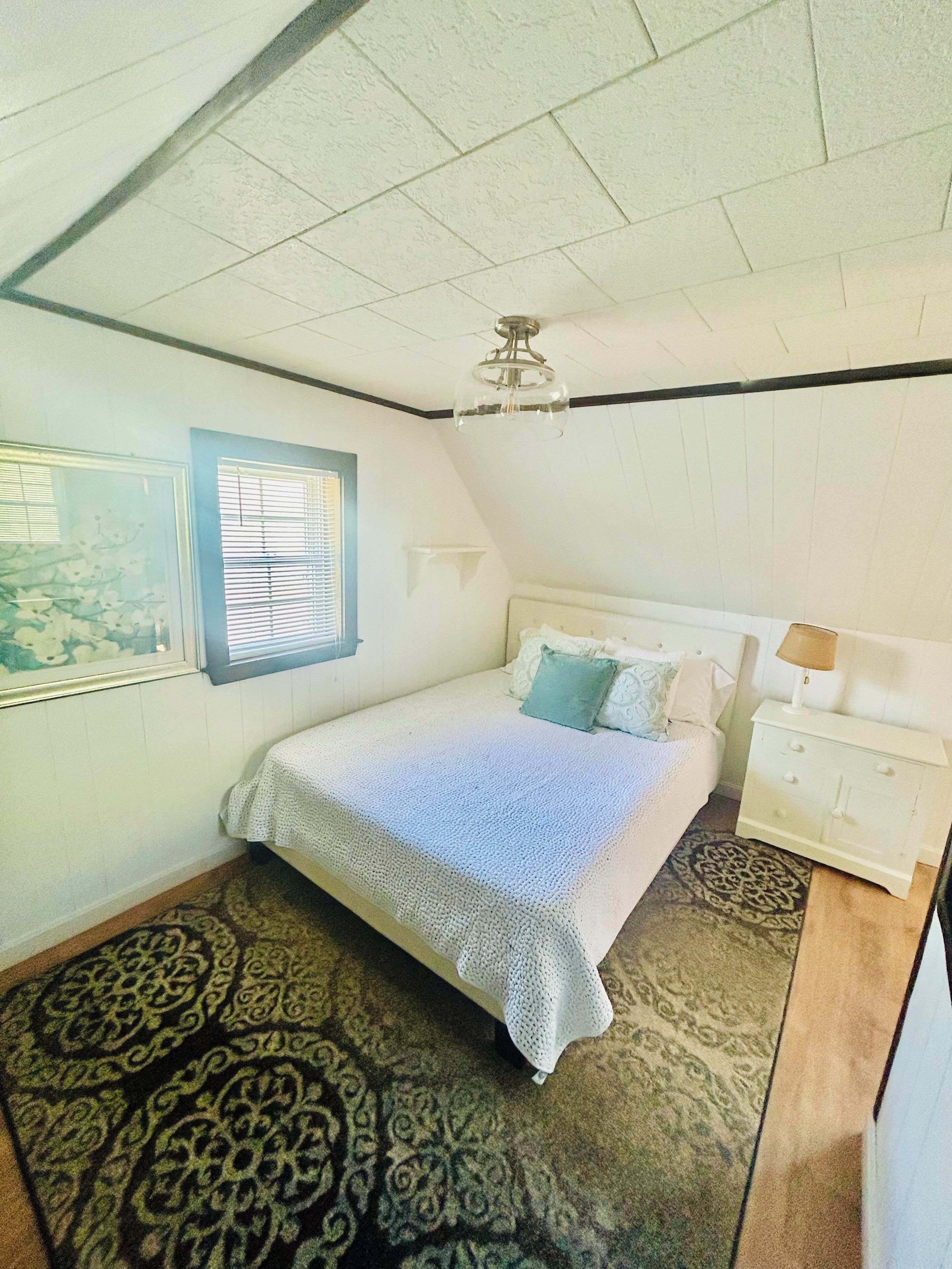 A bedroom with a bed, a nightstand, a lamp, a ceiling fan, a window with blinds, and a patterned rug.