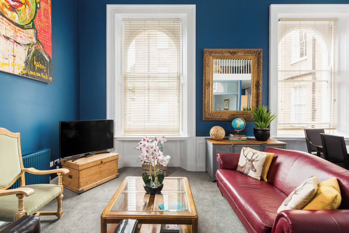 A living room with a red leather sofa, a wooden coffee table with a vase of flowers, a television on a wooden cabinet, and a large mirror on the wall.
