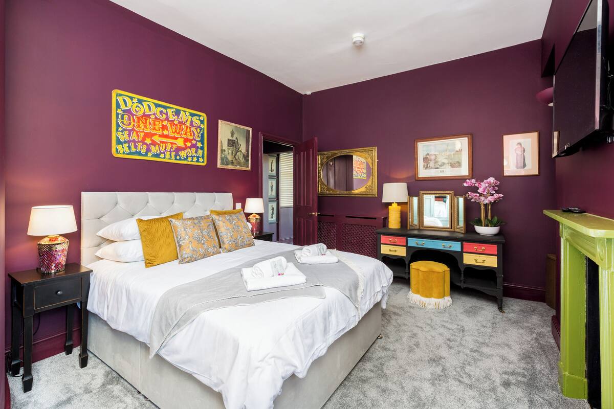 A hotel room with a large bed, a nightstand with a lamp, a dresser with a mirror, and a green fireplace.