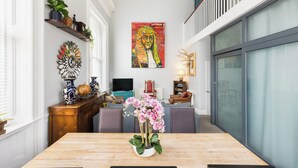 Uma área de refeições com uma mesa de madeira, cadeiras e um vaso de flores cor-de-rosa. Há um quadro na parede e uma televisão sobre um suporte.