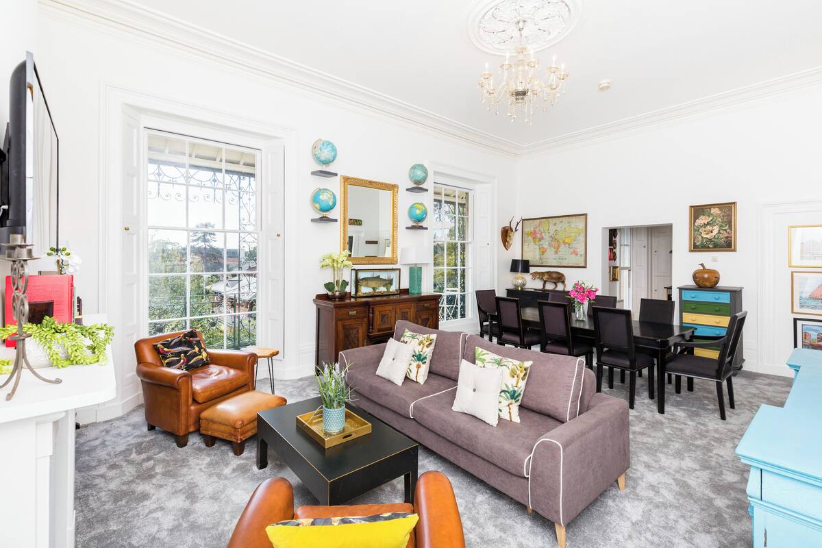 A spacious living room with a brown leather armchair, a gray sofa with white and yellow cushions, and a black coffee table. There is a dining area with a wooden table and chairs, and a blue cabinet.