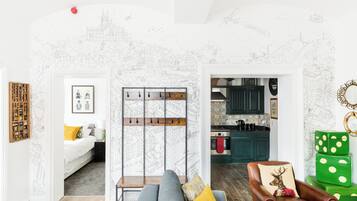 A living room with a grey sofa, a brown leather armchair, and a green polka-dotted ottoman. There is a cityscape mural on the wall, a wooden coat rack, and a framed picture on the wall.