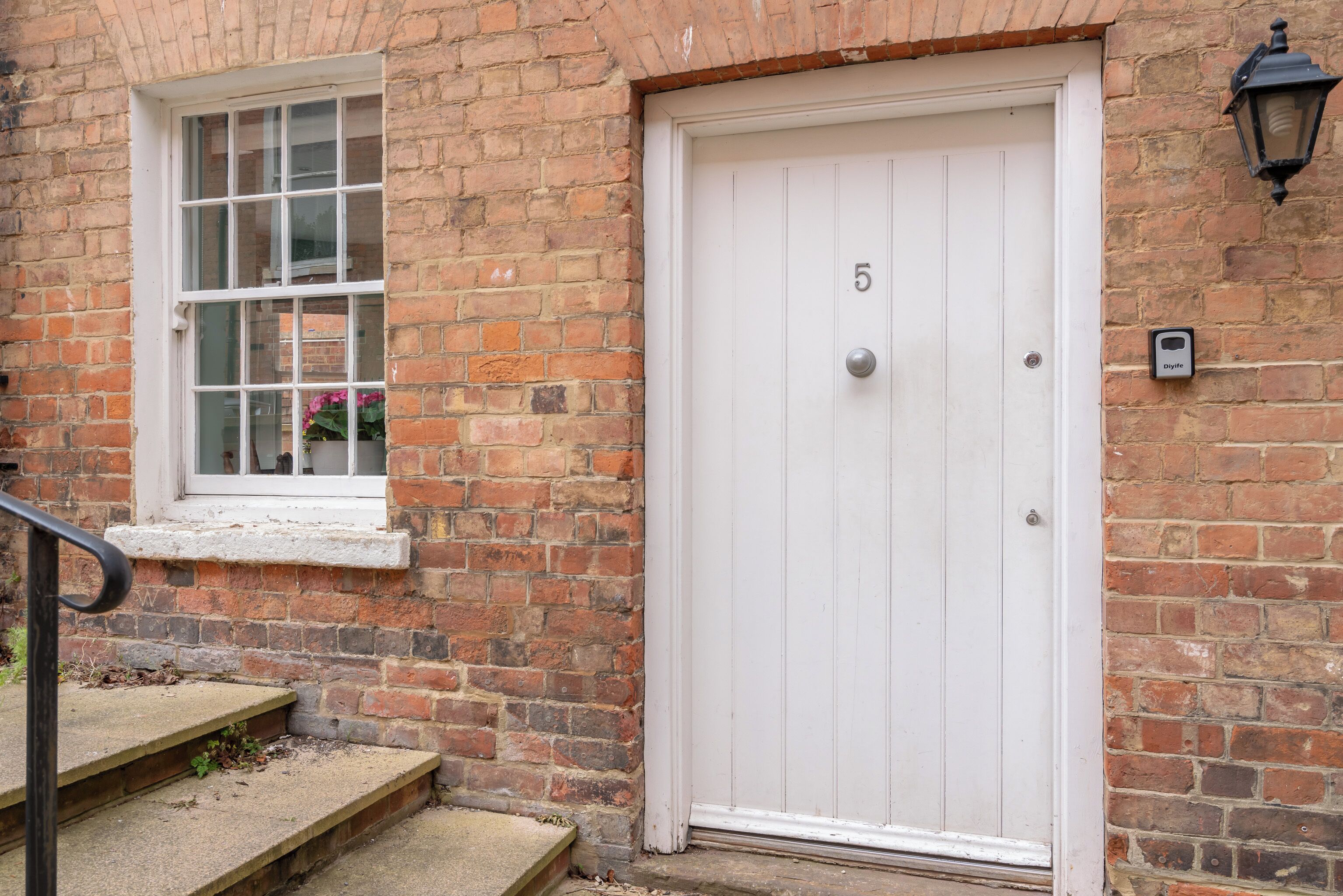 A white door with the number 5, a brick wall, a window with a white frame, and a small step leading to the door.