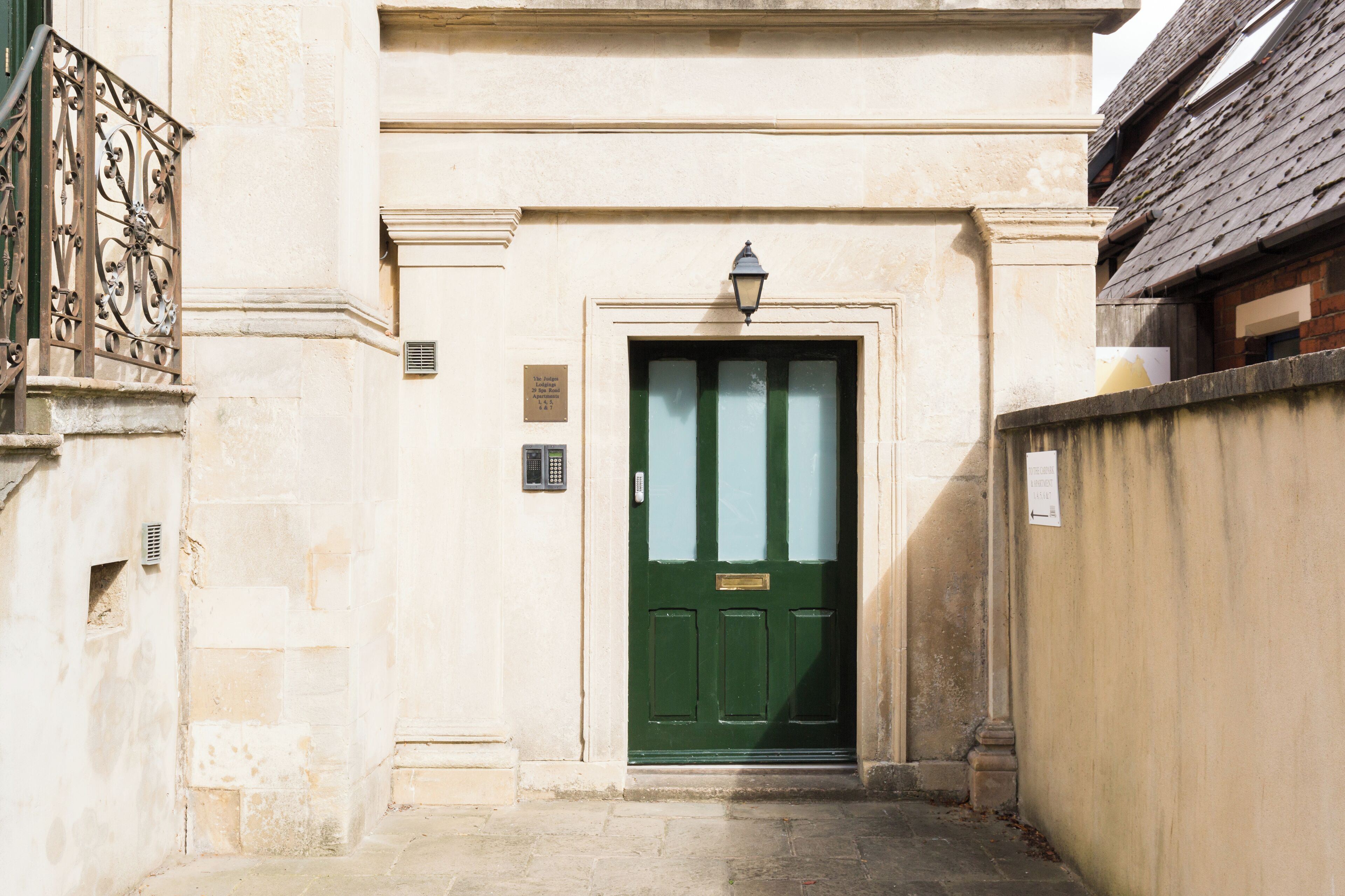 Un edificio con una puerta verde, una ventana pequeña con una lámpara encima y una pared de piedra a la derecha.