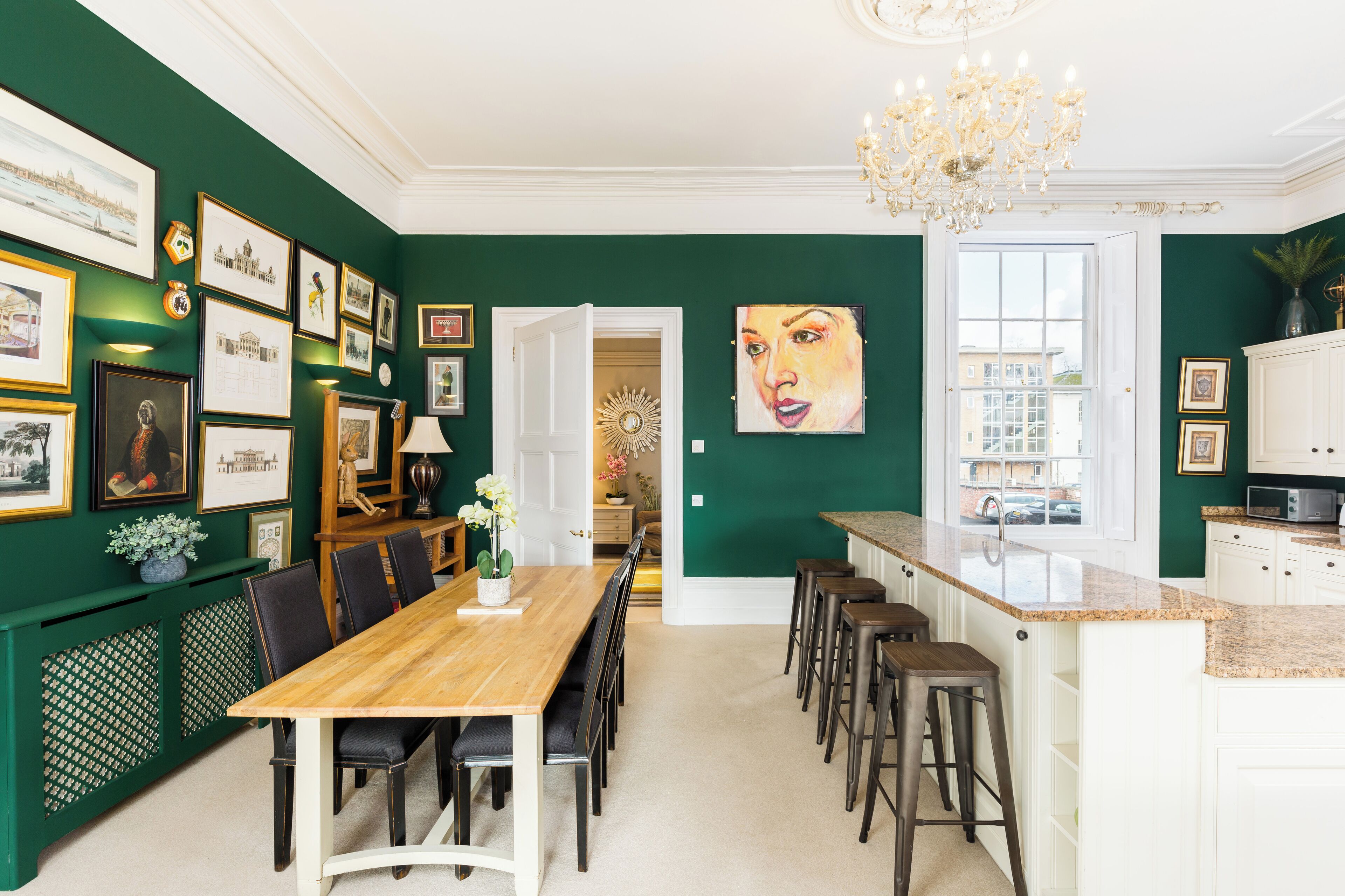 Un comedor con mesa y sillas de madera, una cocina con encimera blanca y una pared verde decorada con marcos.
